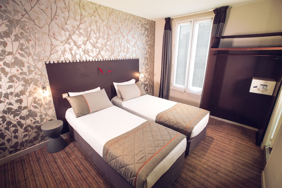 A hotel room with two beds, a nightstand, a wall-mounted heater, and a window with curtains.