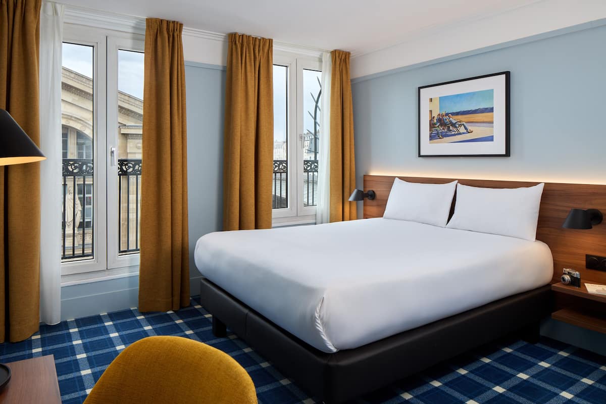 A hotel room with a large bed, a wooden headboard, a nightstand, a framed picture on the wall, and a view of the city through the window.