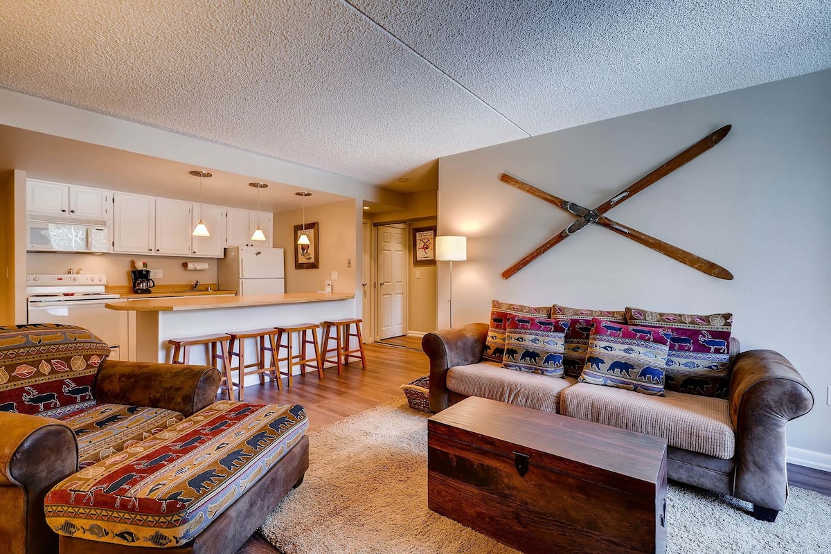 A cozy living room with a sofa, ottoman, and coffee table, a kitchen in the background, and a unique wooden cross on the wall.