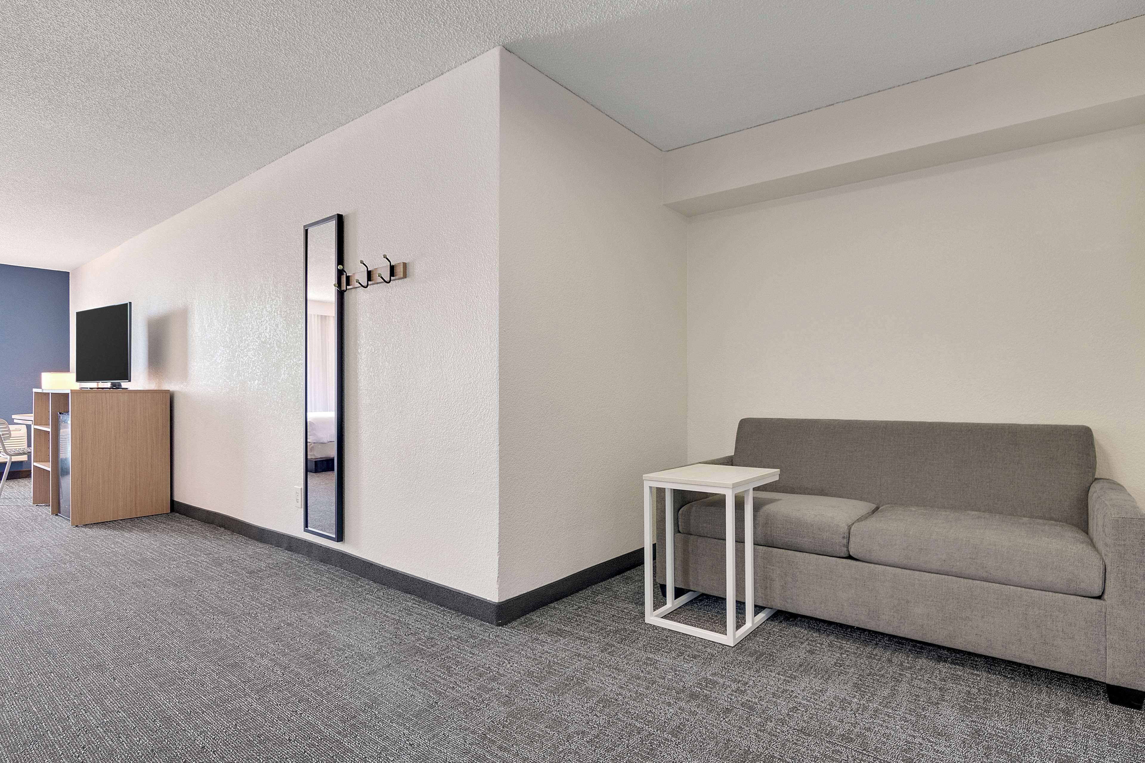 Una habitación de hotel moderna con un sofá gris, una mesita de noche blanca, un espejo grande y un escritorio de madera con televisor de pantalla plana.
