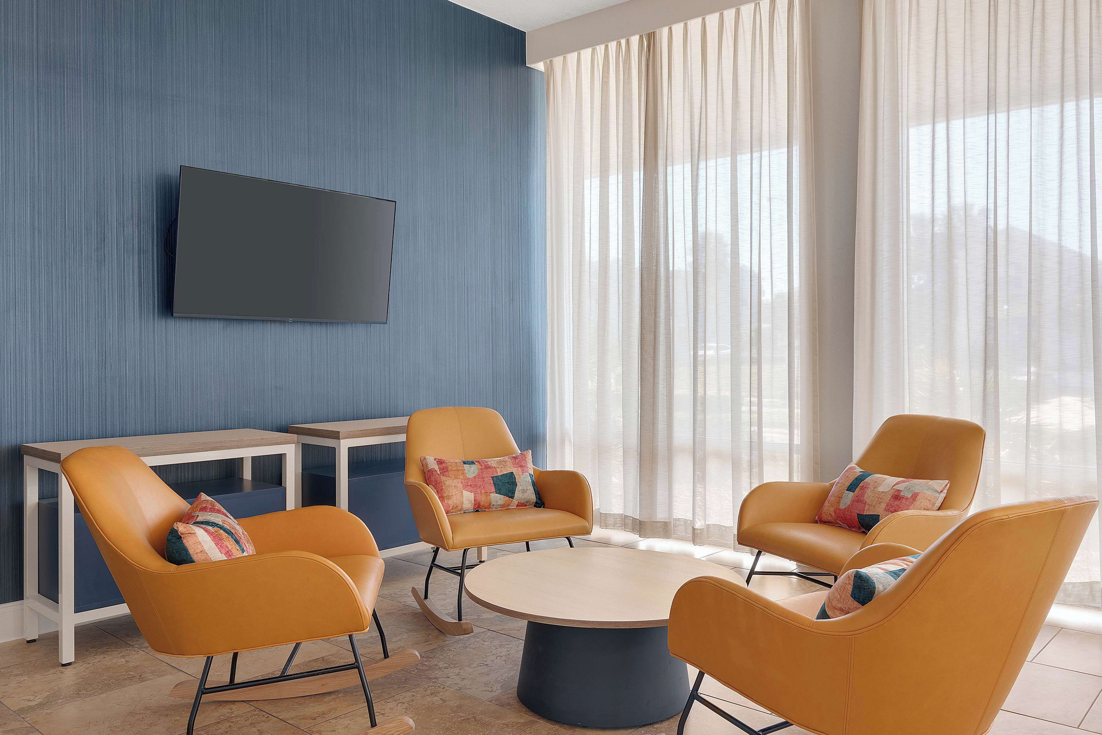 A modern living room with a round coffee table, two orange armchairs, a small white side table, and a flat-screen TV mounted on a blue wall.
