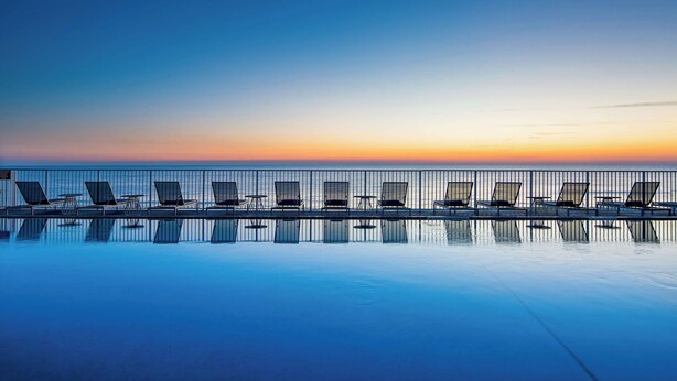 Zona de piscina con sillones de descanso y vista al océano al atardecer.