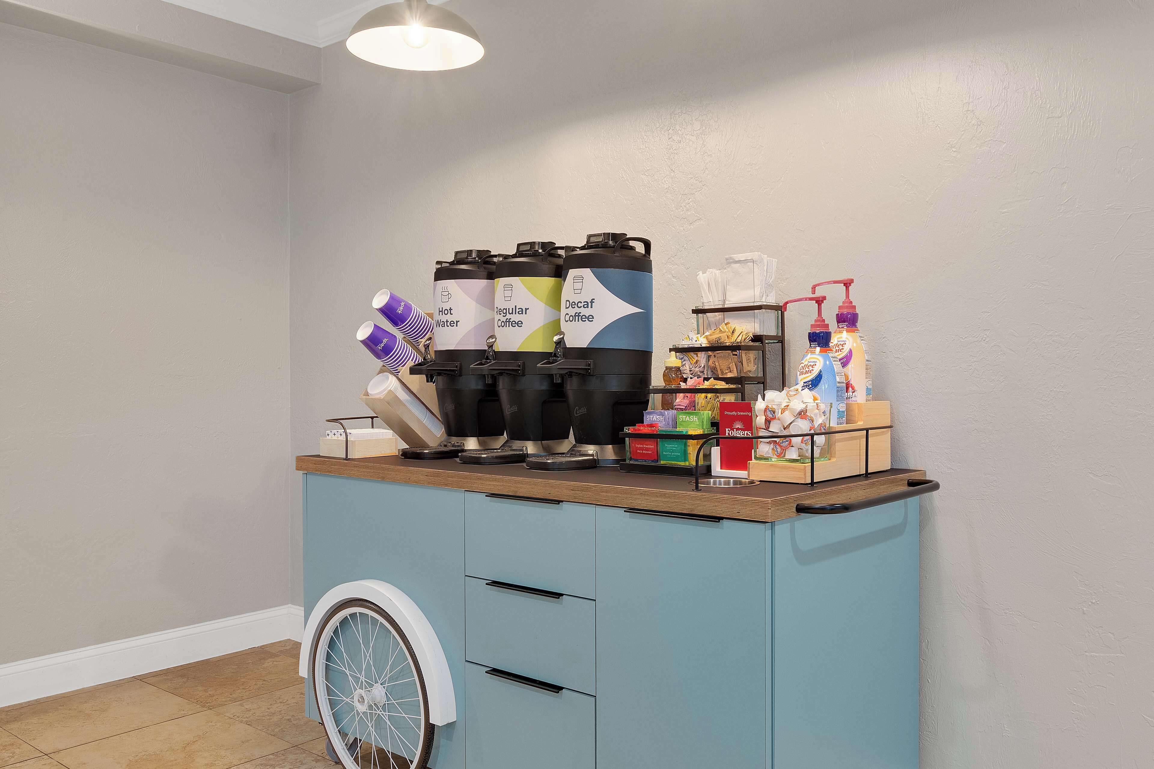 Una estación de café autoservicio con dispensadores etiquetados para agua caliente, café regular y café descafeinado, junto con una variedad de vasos de papel y un dispensador de jabón.