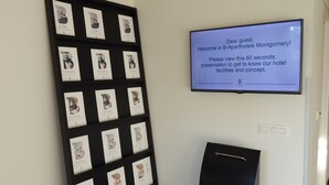 Et hotelreception område med et vægmonteret TV der viser en velkomstbesked. Der er en sort displaystand med indrammede billeder og tekst, samt et sort bagage stativ med oplader.