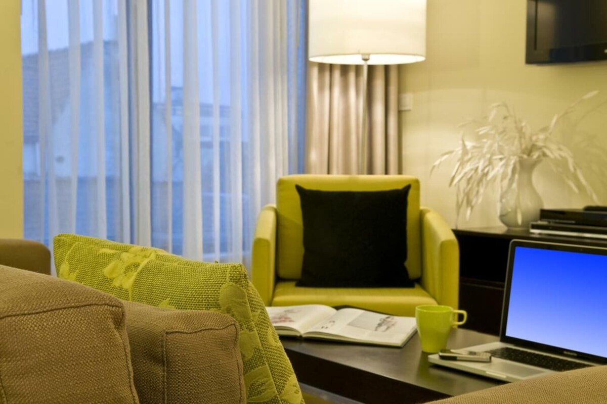 A hotel room with a yellow armchair, a brown sofa, a laptop on a desk, and a window with curtains.
