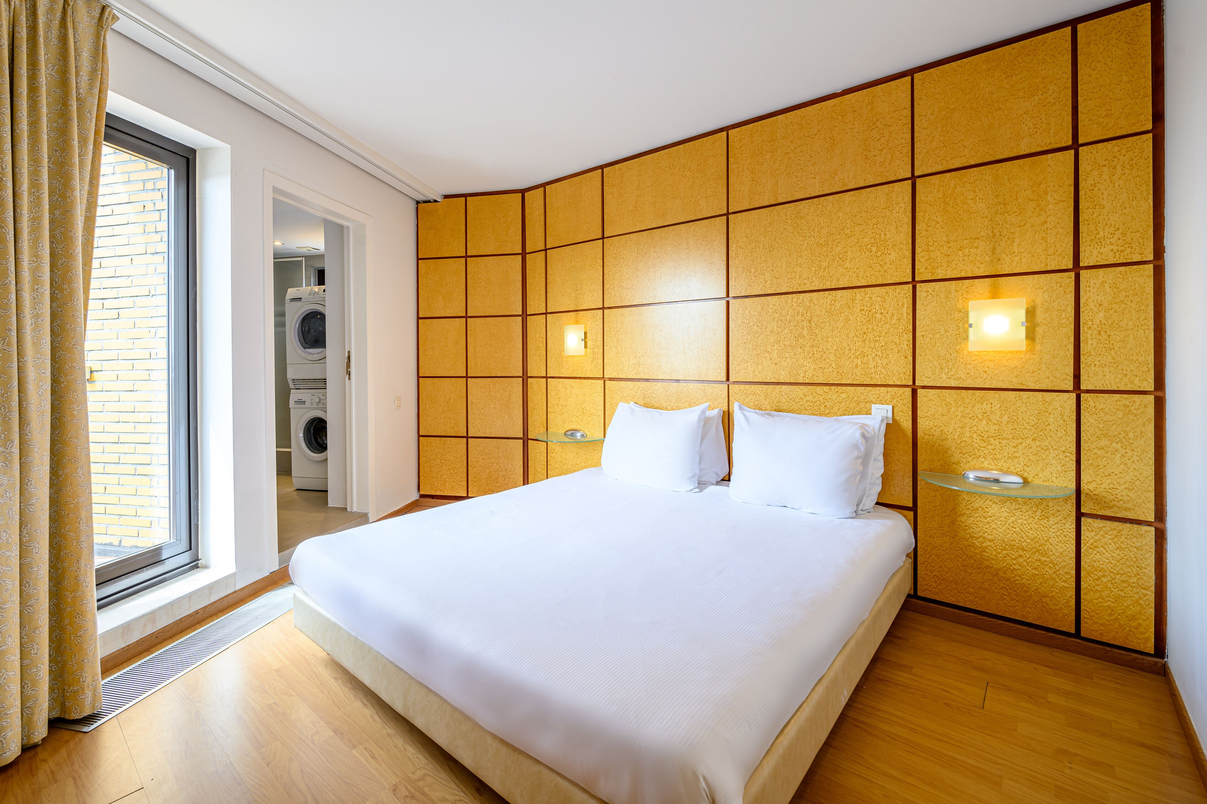 Habitación de hotel con una cama grande, lavadora al fondo y una puerta corrediza de vidrio hacia el exterior.