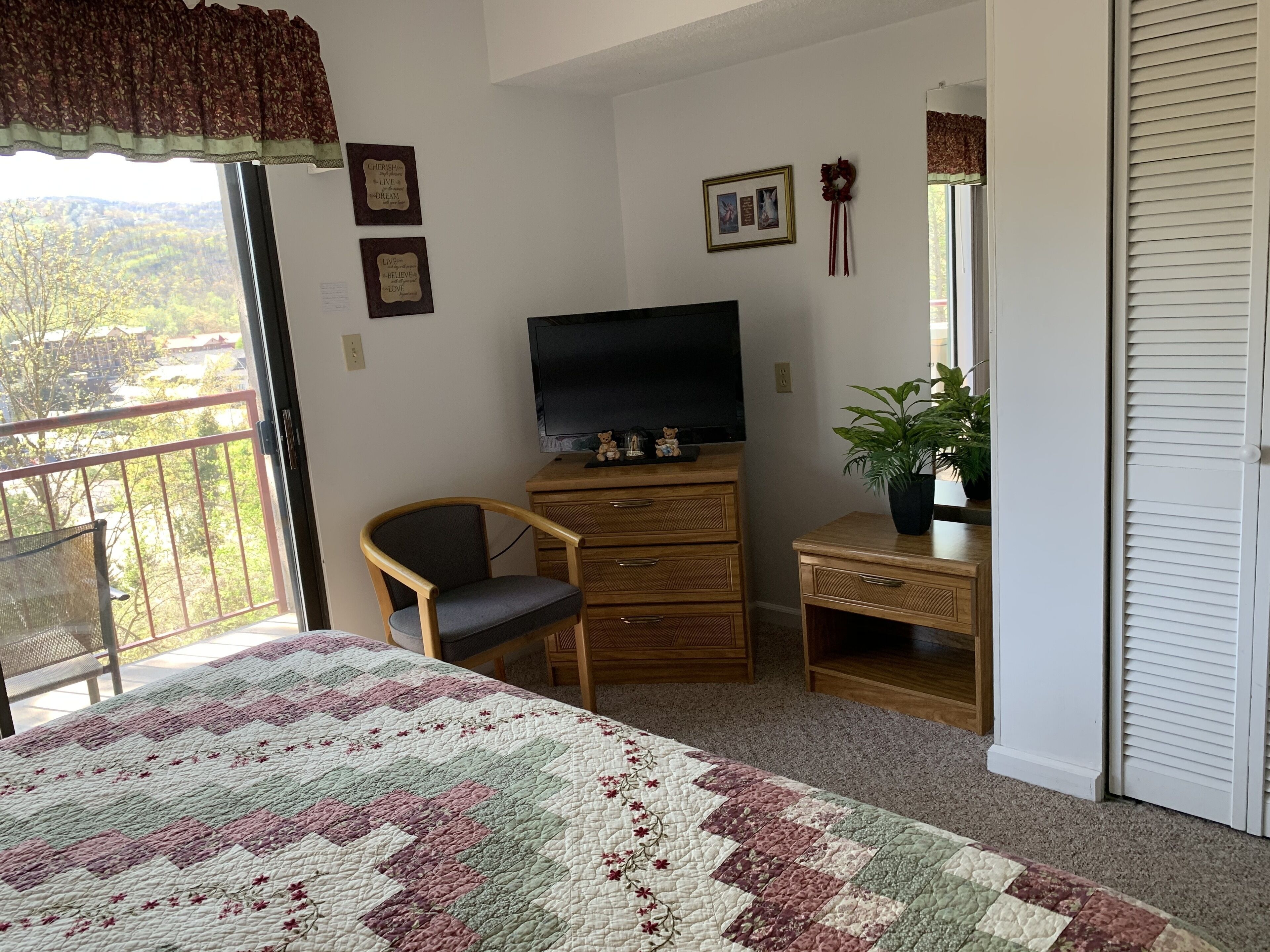 Un dormitorio con cama, televisor, sillón, cómoda y una planta en maceta.