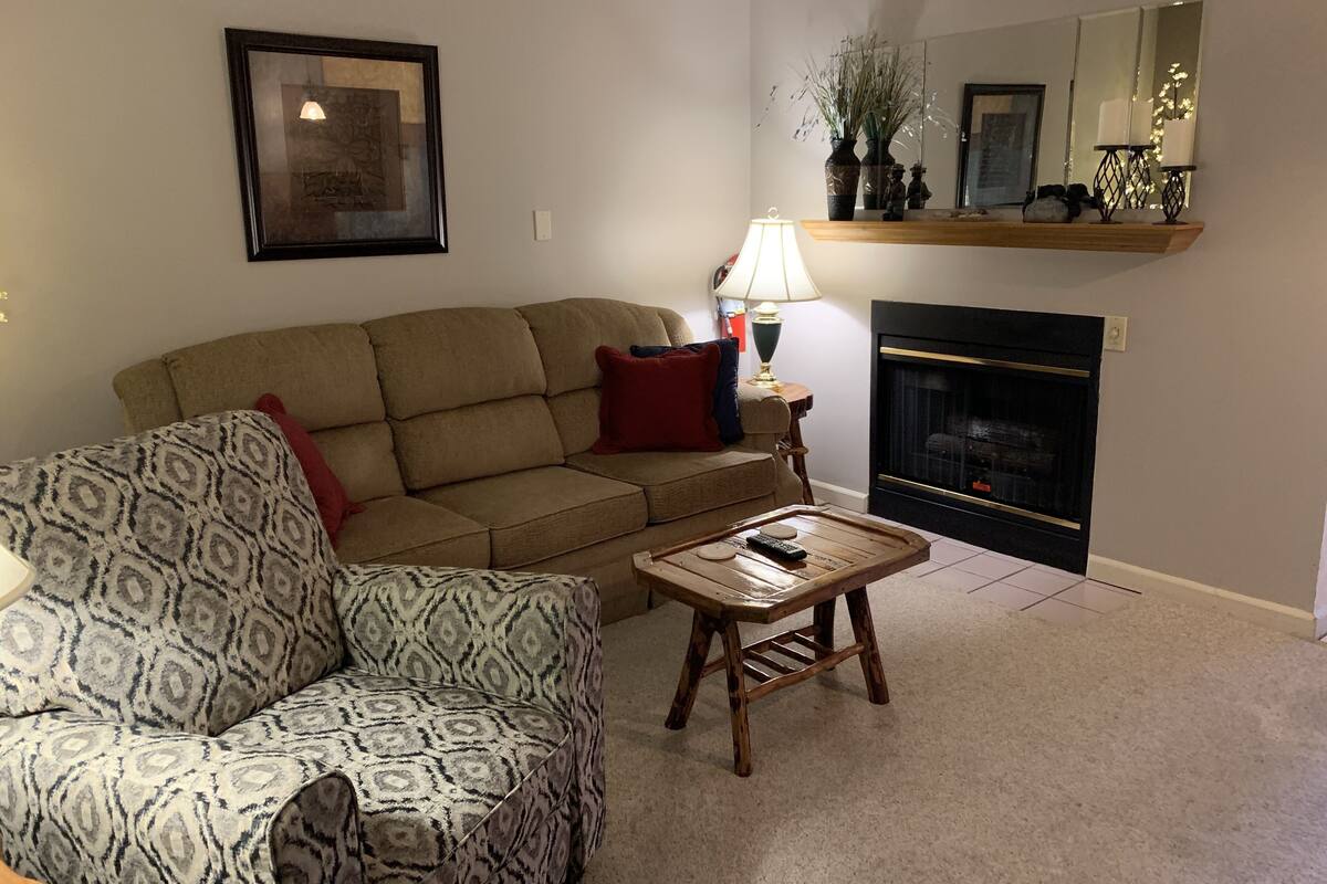 A living room with a sofa, a patterned armchair, a small wooden table, a floor lamp, a fireplace, and a mirror above the mantel.