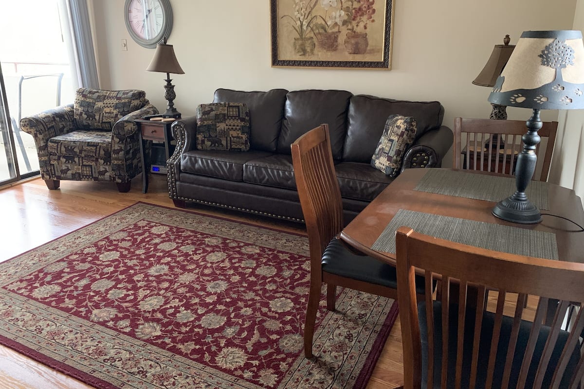 A living room with a dark brown leather sofa, patterned armchairs, a wooden dining table, and a red and beige patterned rug.