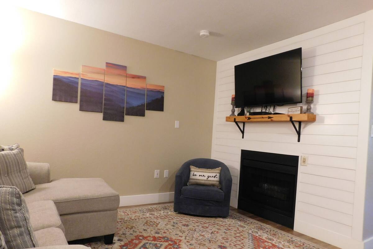 A living room with a beige sofa, a blue armchair, a patterned rug, a fireplace, and a mounted TV.