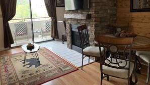 A cozy living room with a stone fireplace, a flat-screen TV mounted on the wall, a dining table with chairs, and a large window with curtains. - Laurel Inn Condominium (Gatlinburg)