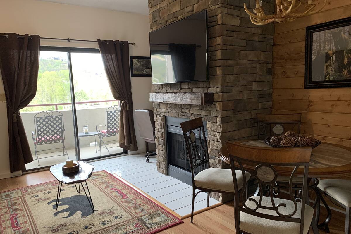 A cozy living room with a stone fireplace, a flat-screen TV mounted on the wall, a dining table with chairs, and a large window with curtains.
