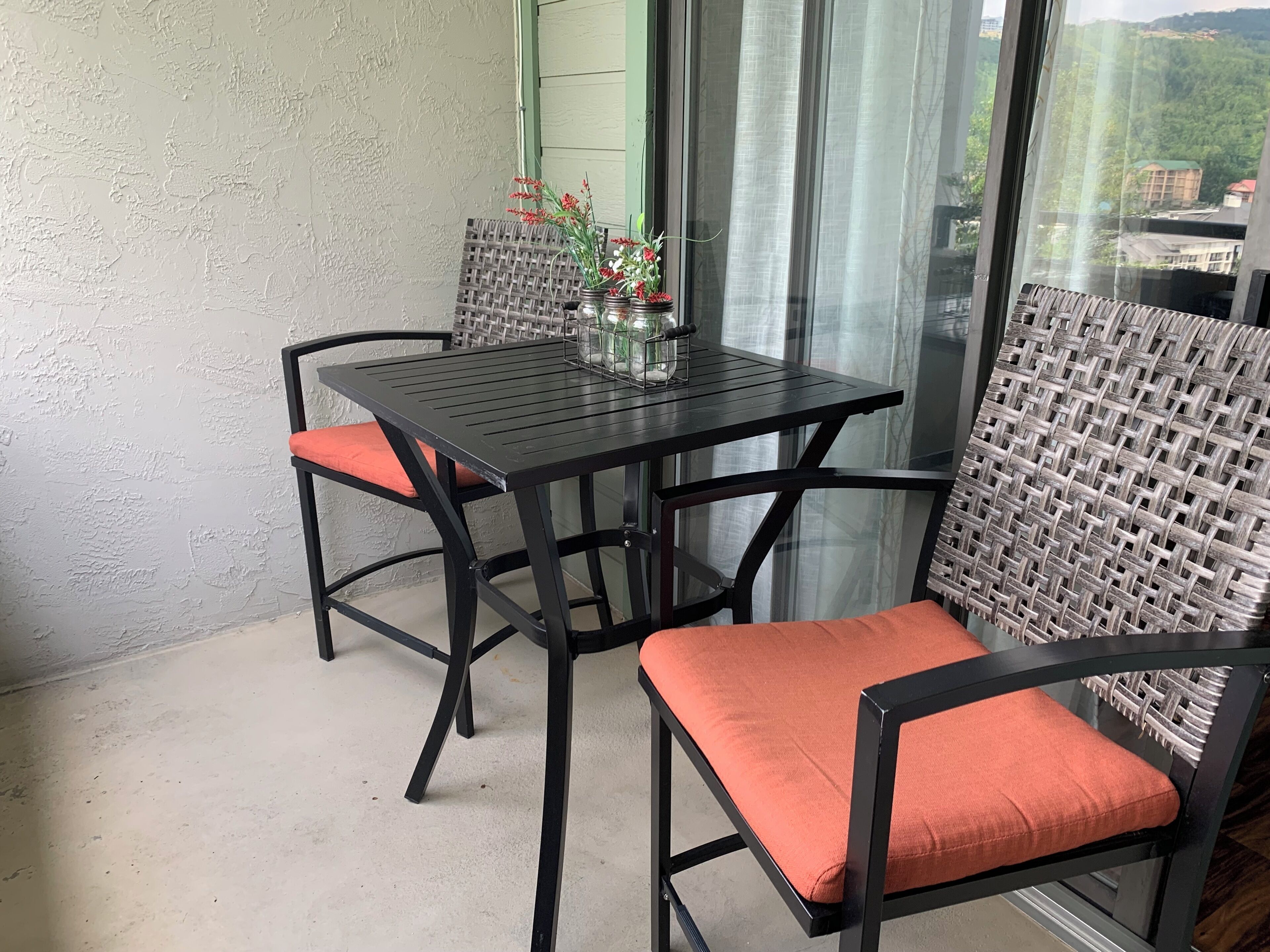 A balcony with a table and chairs, a wicker chair, and a view of buildings through a window.