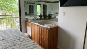 A bedroom with a wooden vanity, a large mirror, a bathtub, and a balcony with a view of trees. - Laurel Inn Condominium (Gatlinburg)