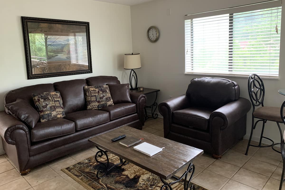 A living room with a brown leather sofa, a matching armchair, a coffee table, a side table with a lamp, and a large window with blinds.