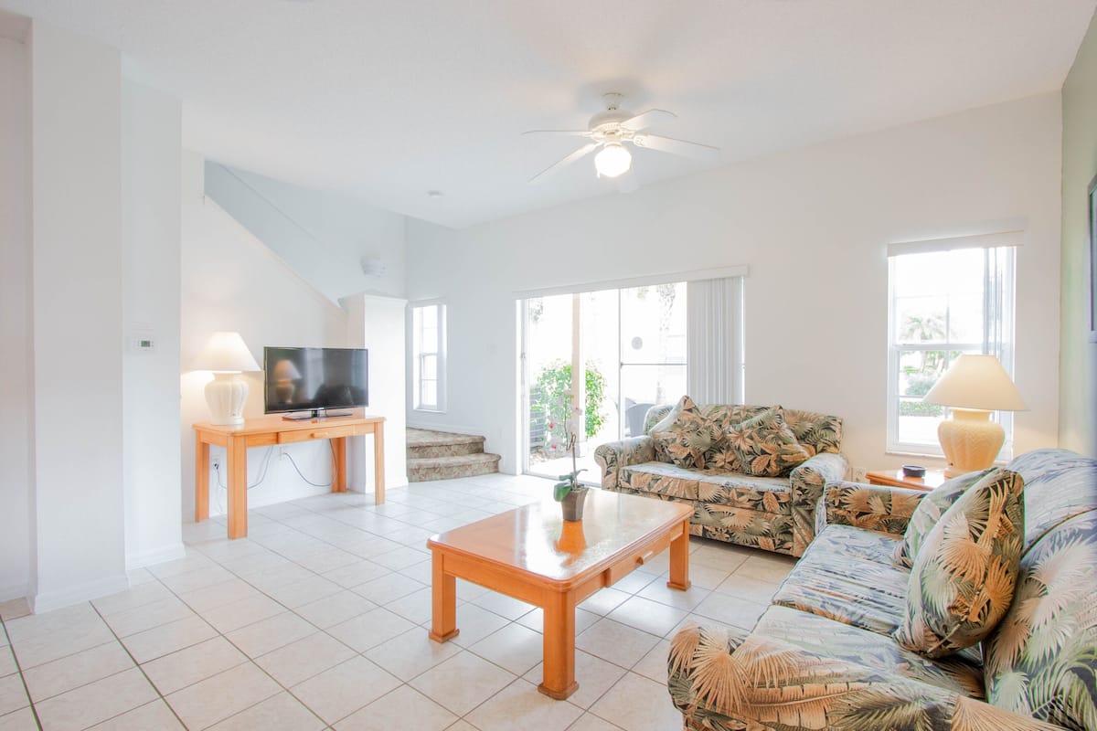 A spacious living room with a television on a wooden stand, a sofa with a tropical print, and a ceiling fan.