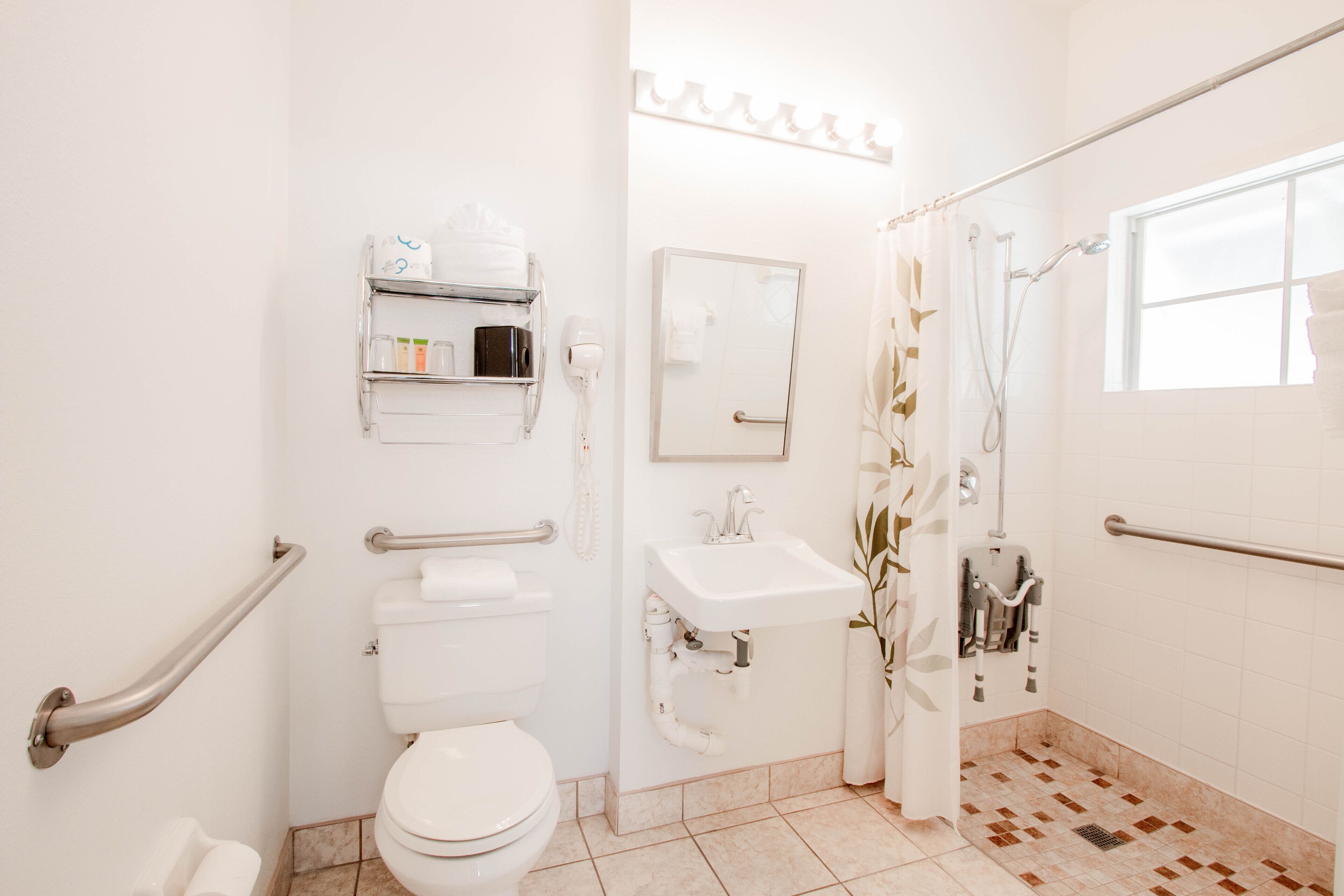 Un baño con inodoro, lavamanos y ducha. Hay barras de apoyo y un espejo.