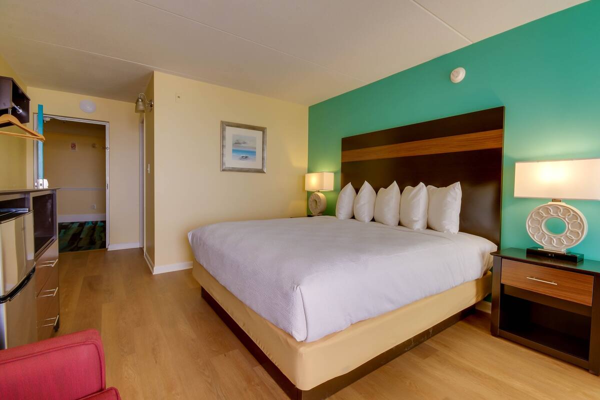 A hotel room with a large bed, wooden flooring, a desk with a lamp, and a view of the sea through the window.
