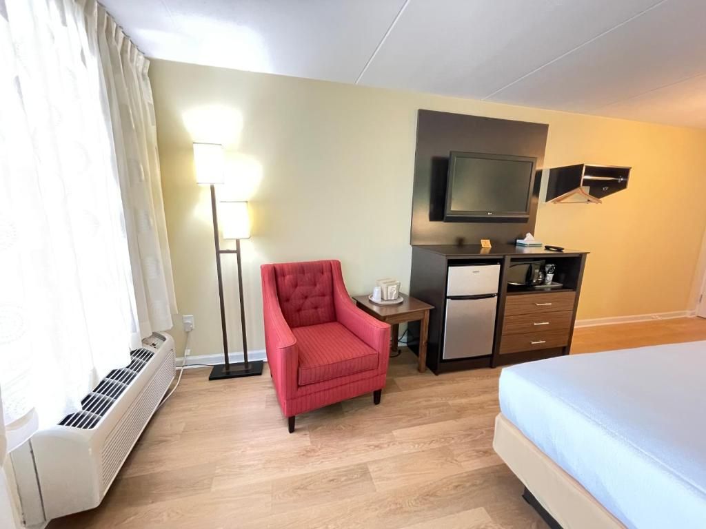 Une chambre d’hôtel comprenant un lit, un fauteuil rouge, un bureau avec un téléviseur à écran plat, un mini-réfrigérateur et un radiateur.