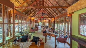 An indoor dining area with wooden tables and chairs, large windows, and a thatched roof ceiling.