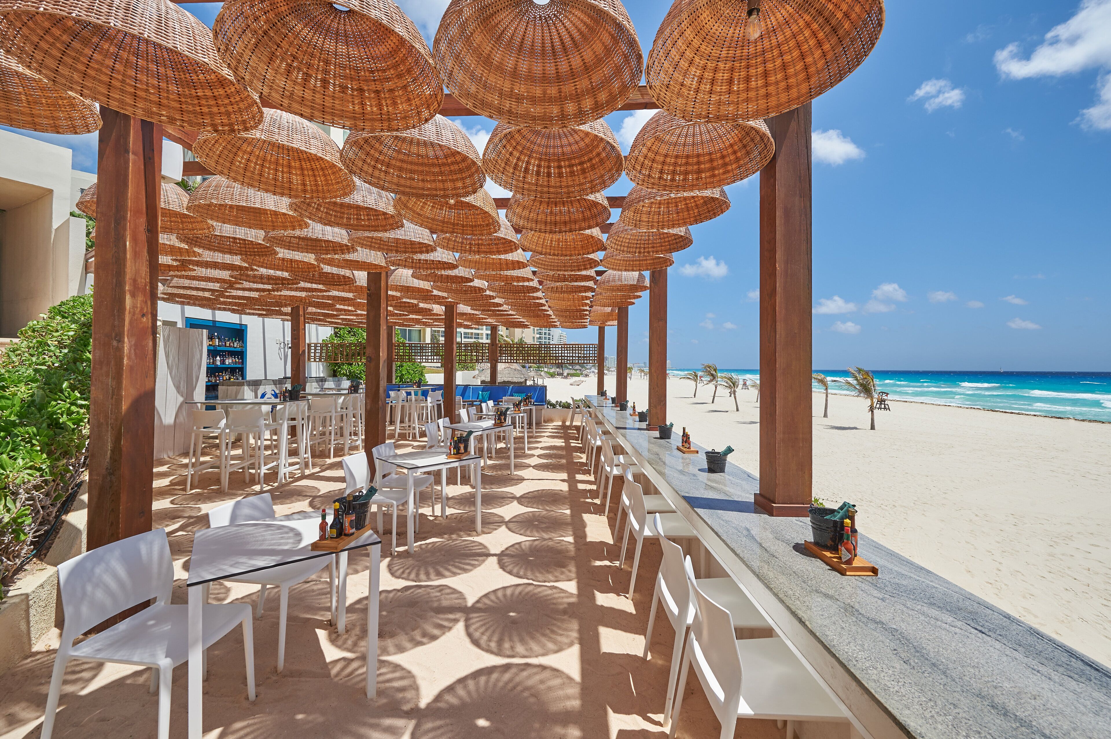 An outdoor dining area with white chairs and tables, large woven umbrellas, and a view of the beach and ocean.