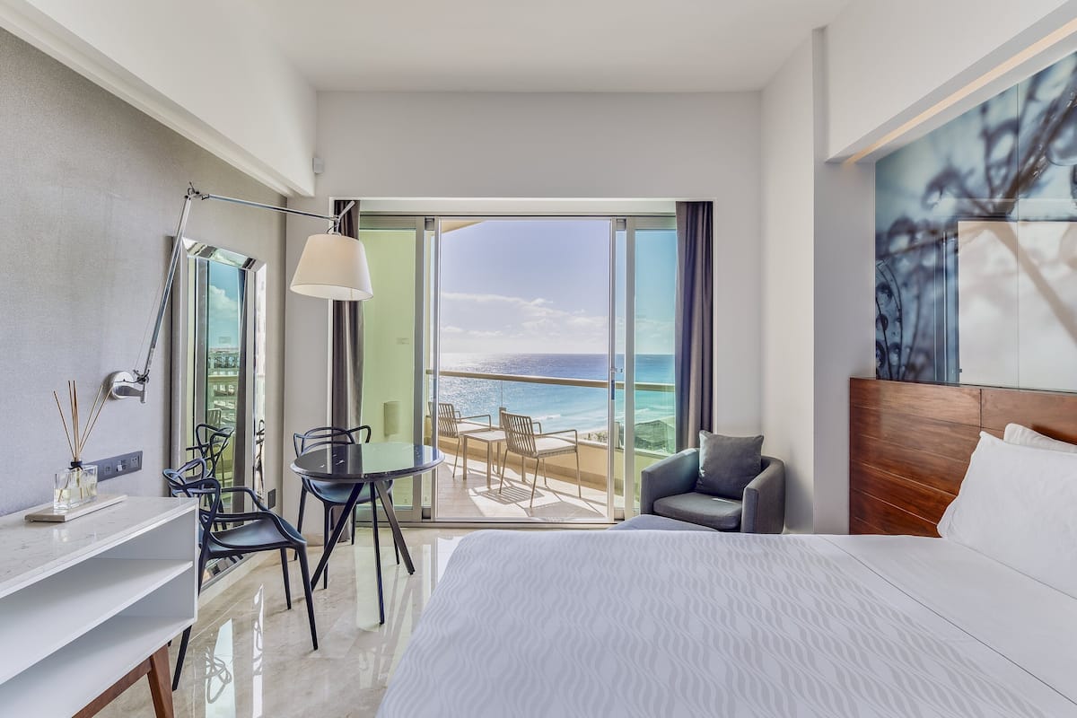Ein modernes Hotelzimmer mit einem großen Bett, einem Schreibtisch, einem Sessel und einem Balkon mit Meerblick.