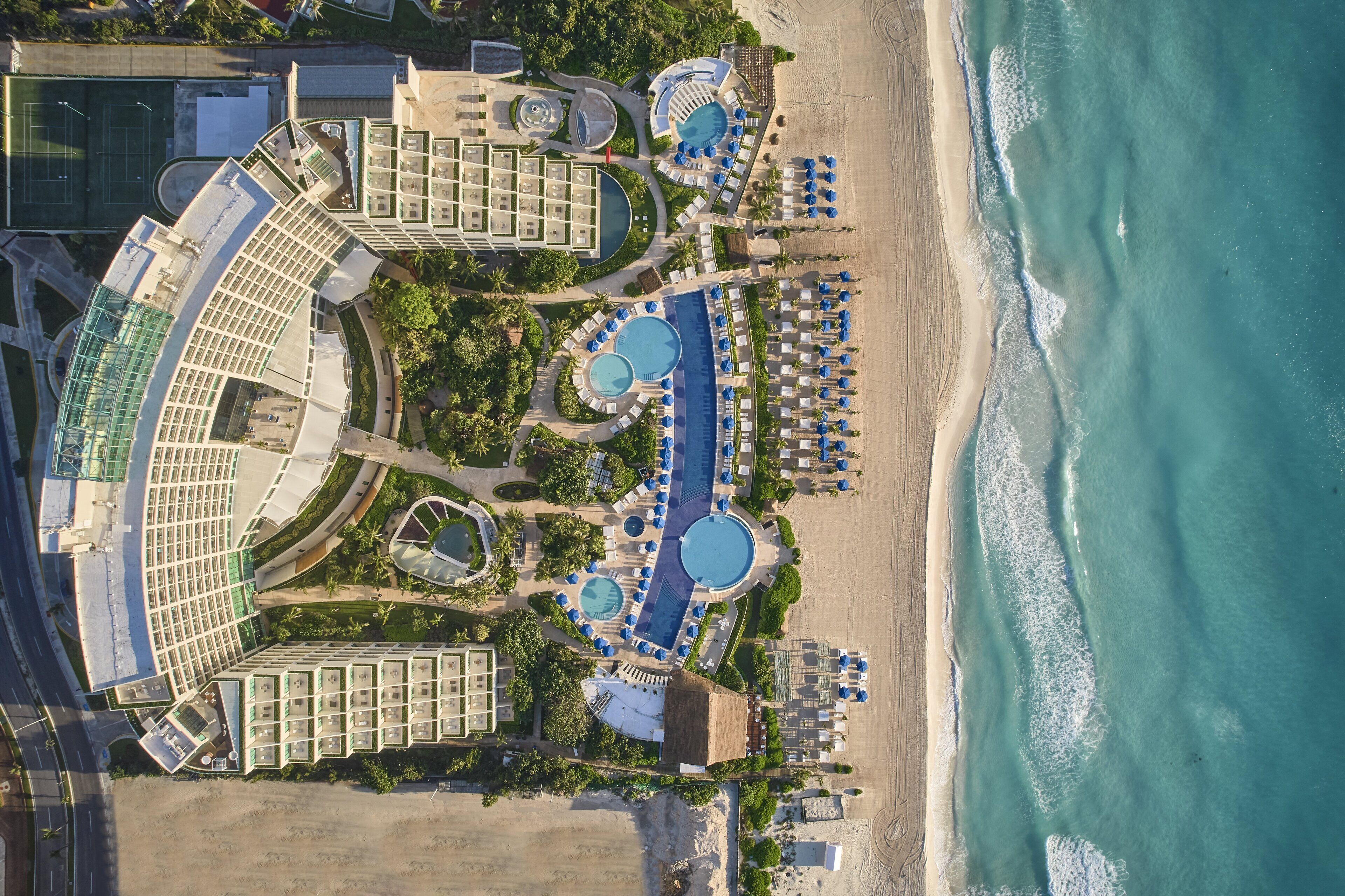 An aerial view of a resort with multiple swimming pools, beachside lounge areas, and a large building complex.
