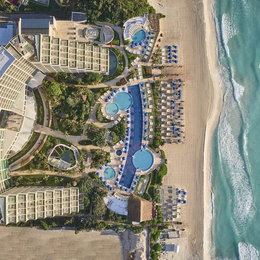 An aerial view of a resort with multiple swimming pools, beachside lounge areas, and a large building complex.