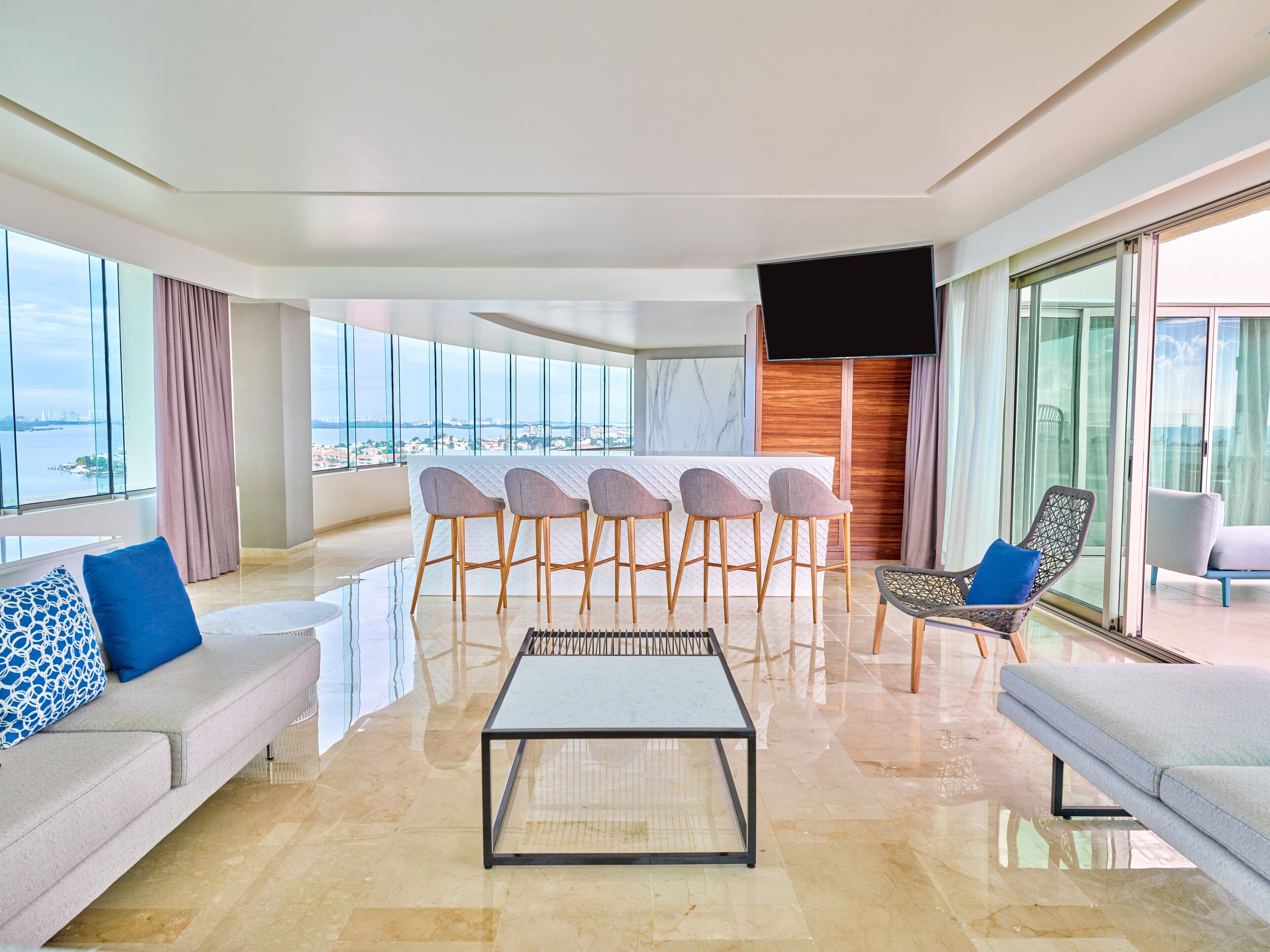 A modern living room with a sofa, a coffee table, and a bar area with stools. There is a large TV mounted on the wall and a view of the city through large windows.