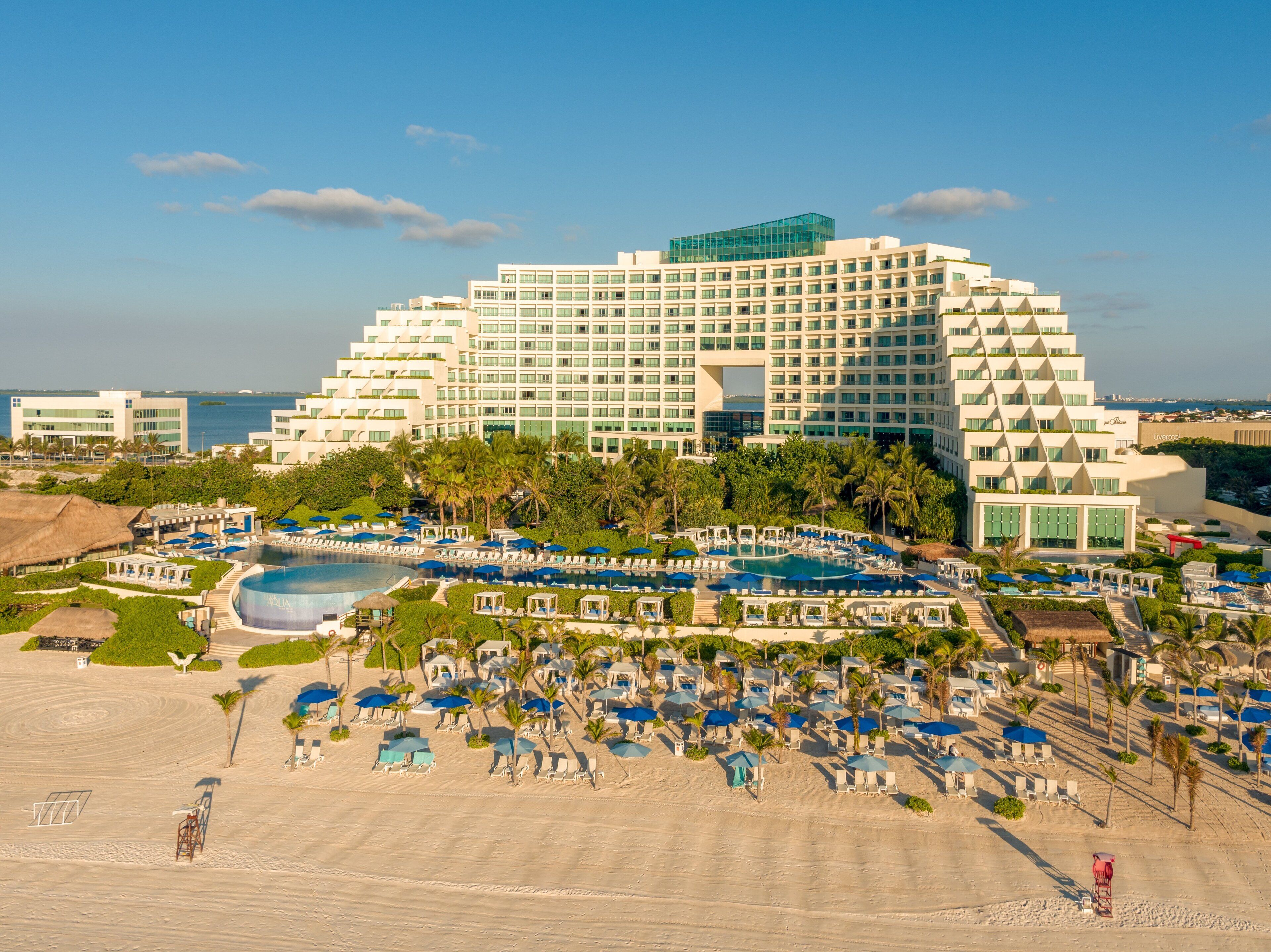 Een groot hotel met meerdere zwembaden, een strandgedeelte met parasols en uitzicht op zee.
