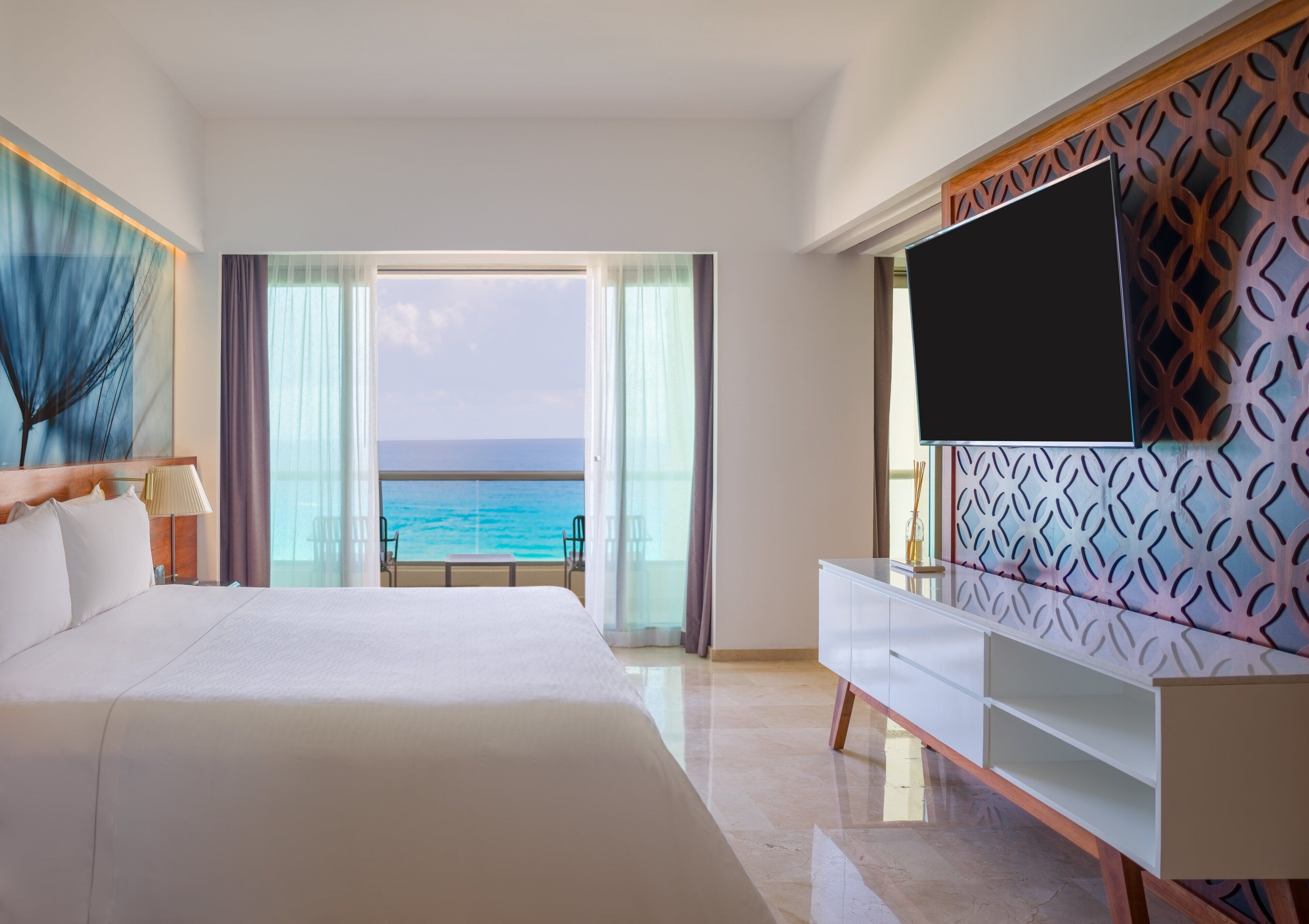 A hotel room with a large bed, a TV mounted on a decorative wall, and a view of the ocean through a large window.