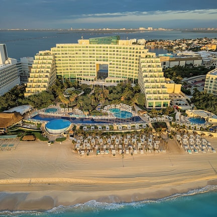 A large hotel with multiple swimming pools, a beach area with sun loungers, and a view of the ocean.