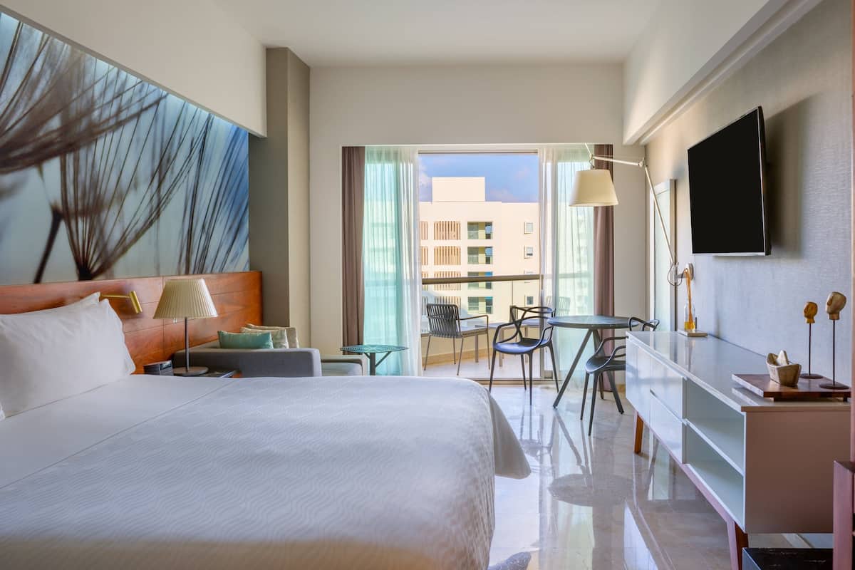 Ein Hotelzimmer mit einem großen Bett, einem Schreibtisch, einem Stuhl und einem Balkon mit Blick auf Gebäude.