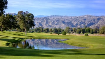 Un campo de golf con un estanque, árboles y montañas al fondo.