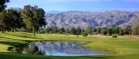 Un parcours de golf avec un étang, des arbres et des montagnes en arrière-plan.