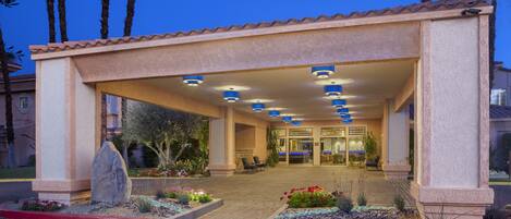 A hotel entrance with a covered walkway, palm trees, and a well-maintained garden.