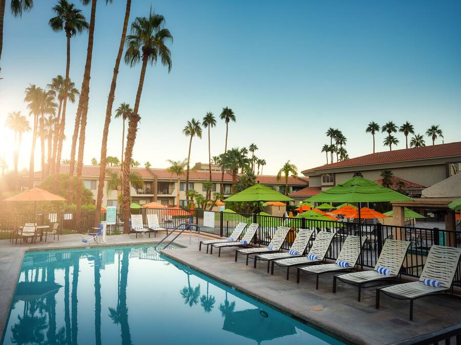 A pool area with lounge chairs and umbrellas, surrounded by palm trees and hotel buildings.