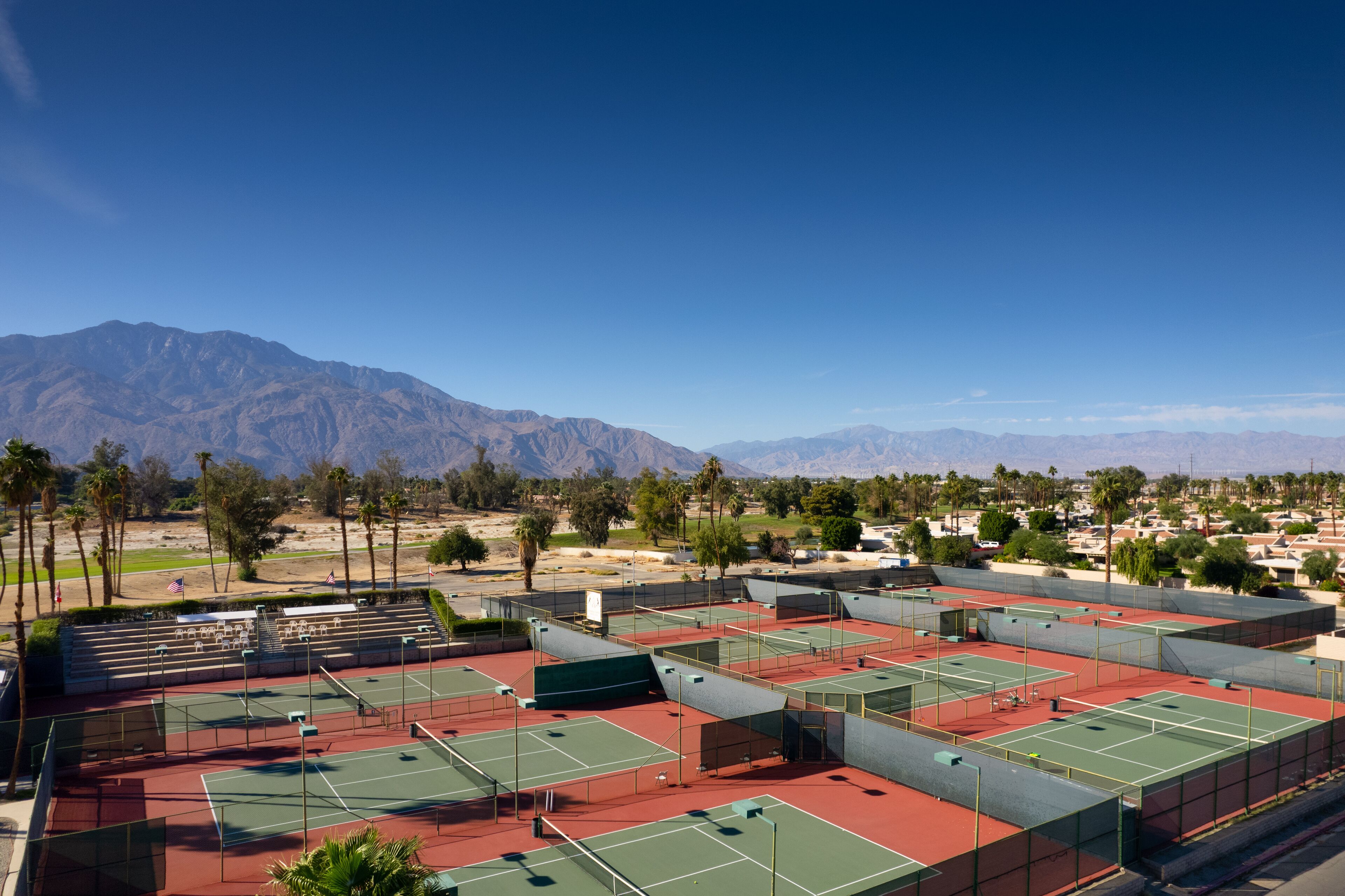 A tennis court complex with multiple courts, seating areas, and a backdrop of mountains.