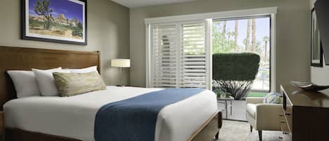 A hotel room with a large bed, a ceiling fan, a desk with a chair, a window with blinds, and a framed picture on the wall.