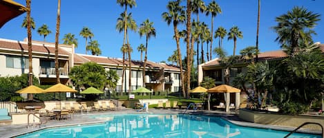 Un área de piscina con sillones de descanso y sombrillas, rodeada de altas palmeras y un edificio con balcones.