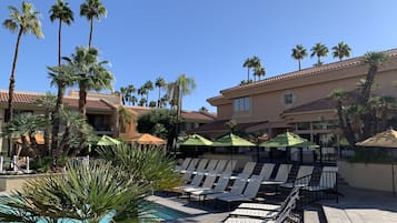 A poolside area with lounge chairs and umbrellas, surrounded by palm trees and a building with a tiled roof.