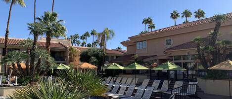 A poolside area with lounge chairs and umbrellas, surrounded by palm trees and a building with a tiled roof.