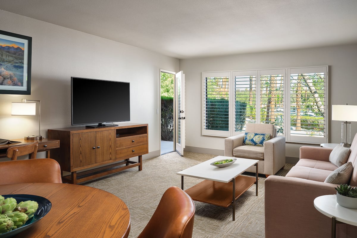 A modern living room with a flat-screen TV, wooden furniture, a dining table with a bowl of fruit, and large windows with blinds.