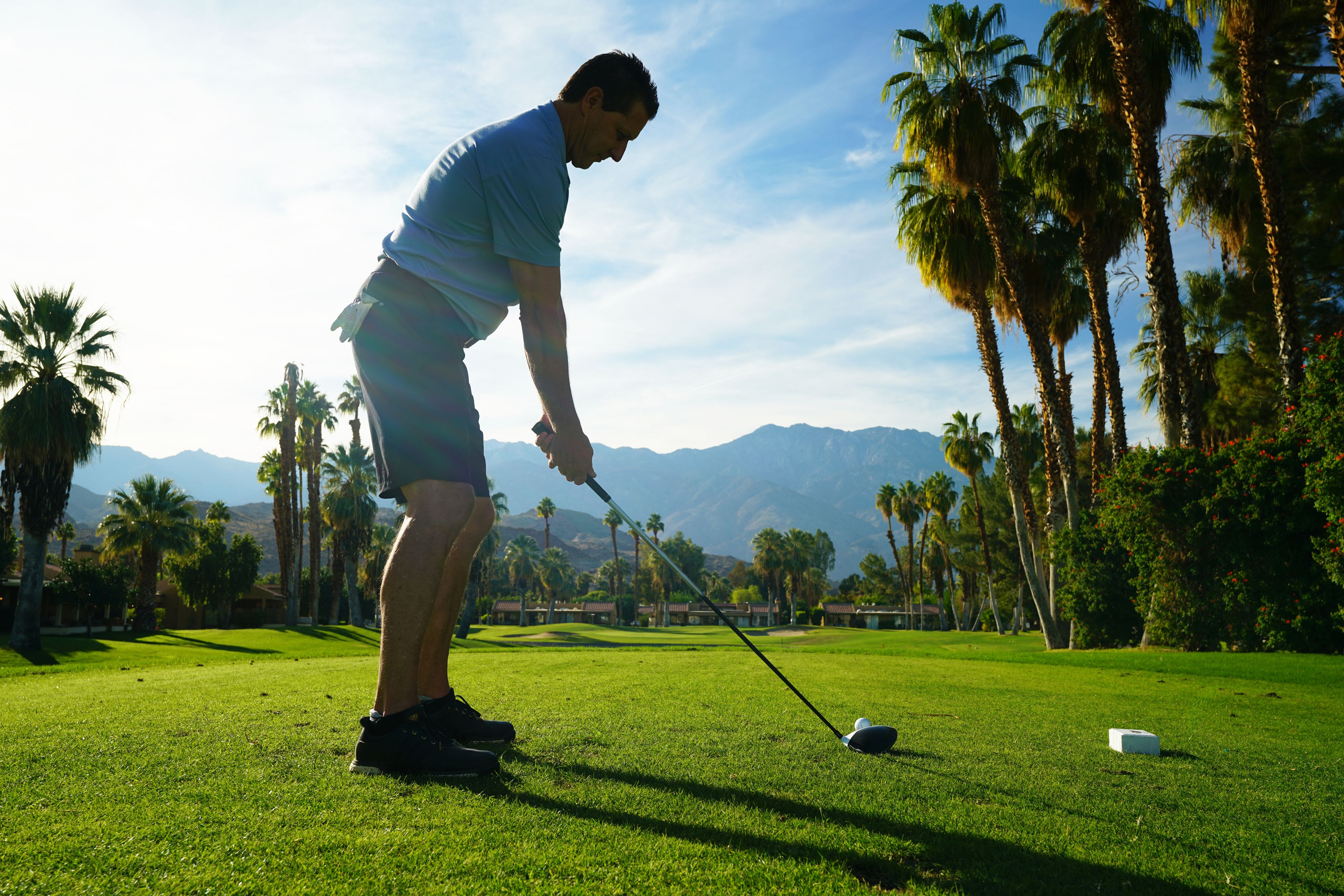 A person on a golf course with palm trees and mountains in the background.