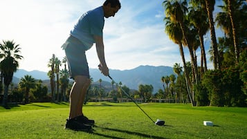 Una persona en un campo de golf, con palmeras y montañas al fondo.