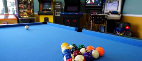 A pool table with a set of billiard balls and arcade games in the background.
