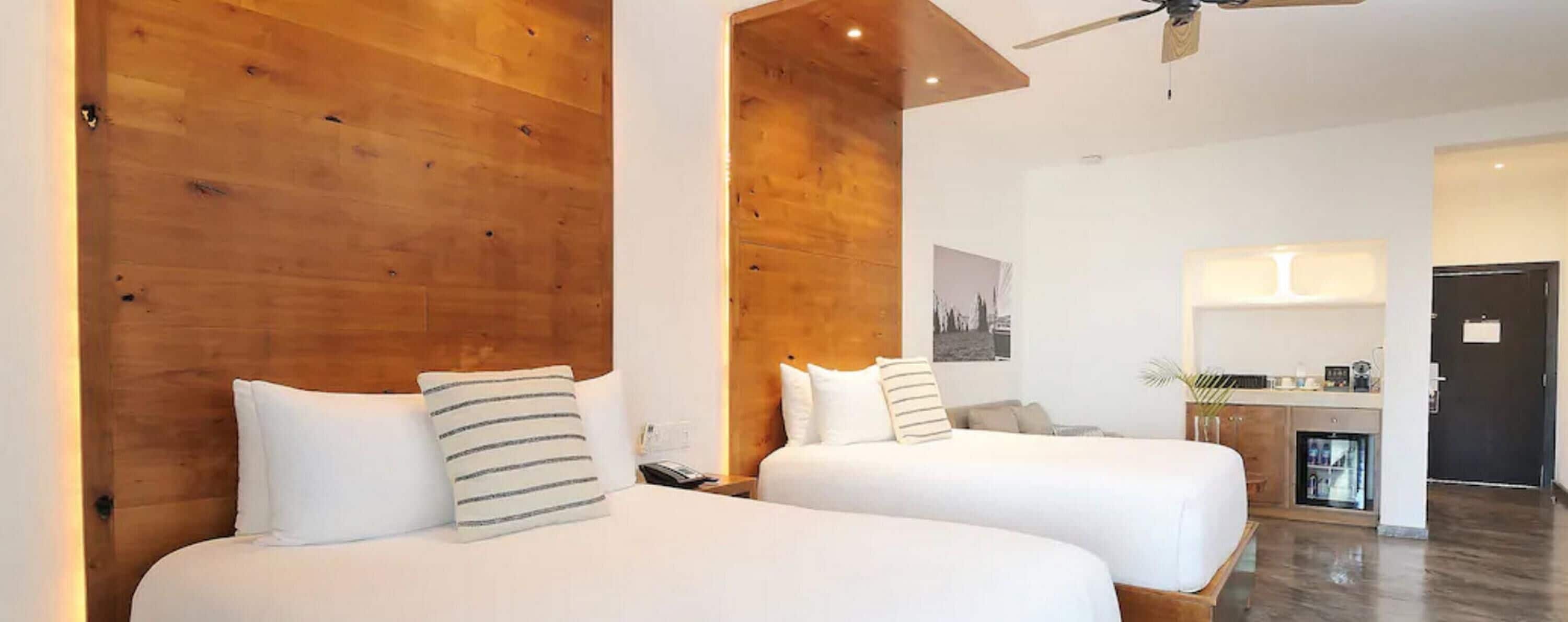 Une chambre d’hôtel avec deux lits, des têtes de lit en bois, un ventilateur de plafond et, au fond, une petite cuisine.