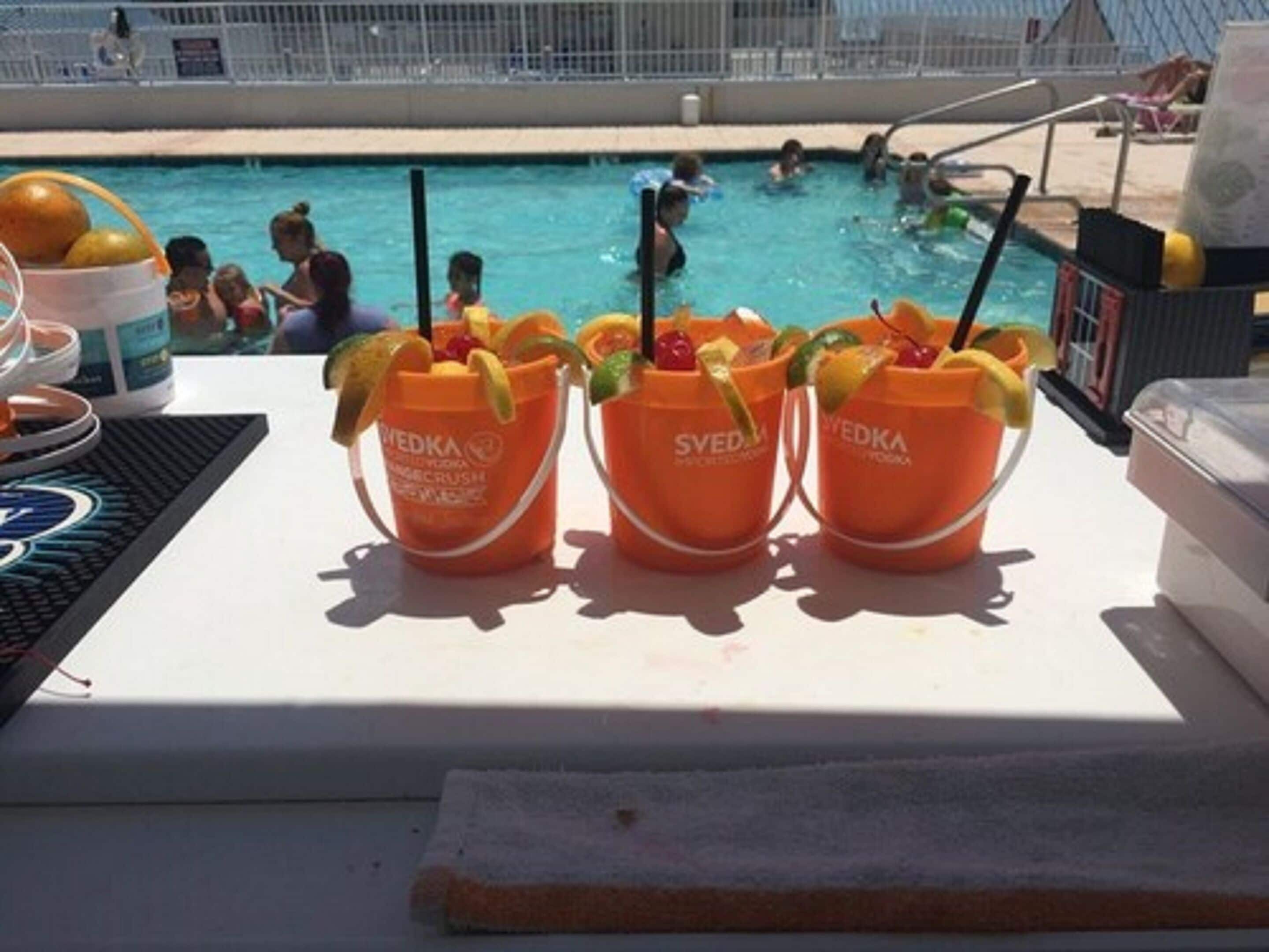 Une table placée au bord de la piscine, sur laquelle se trouvent trois seaux orange de la marque « Svedka », remplis de boissons décorées de fruits et de pailles.