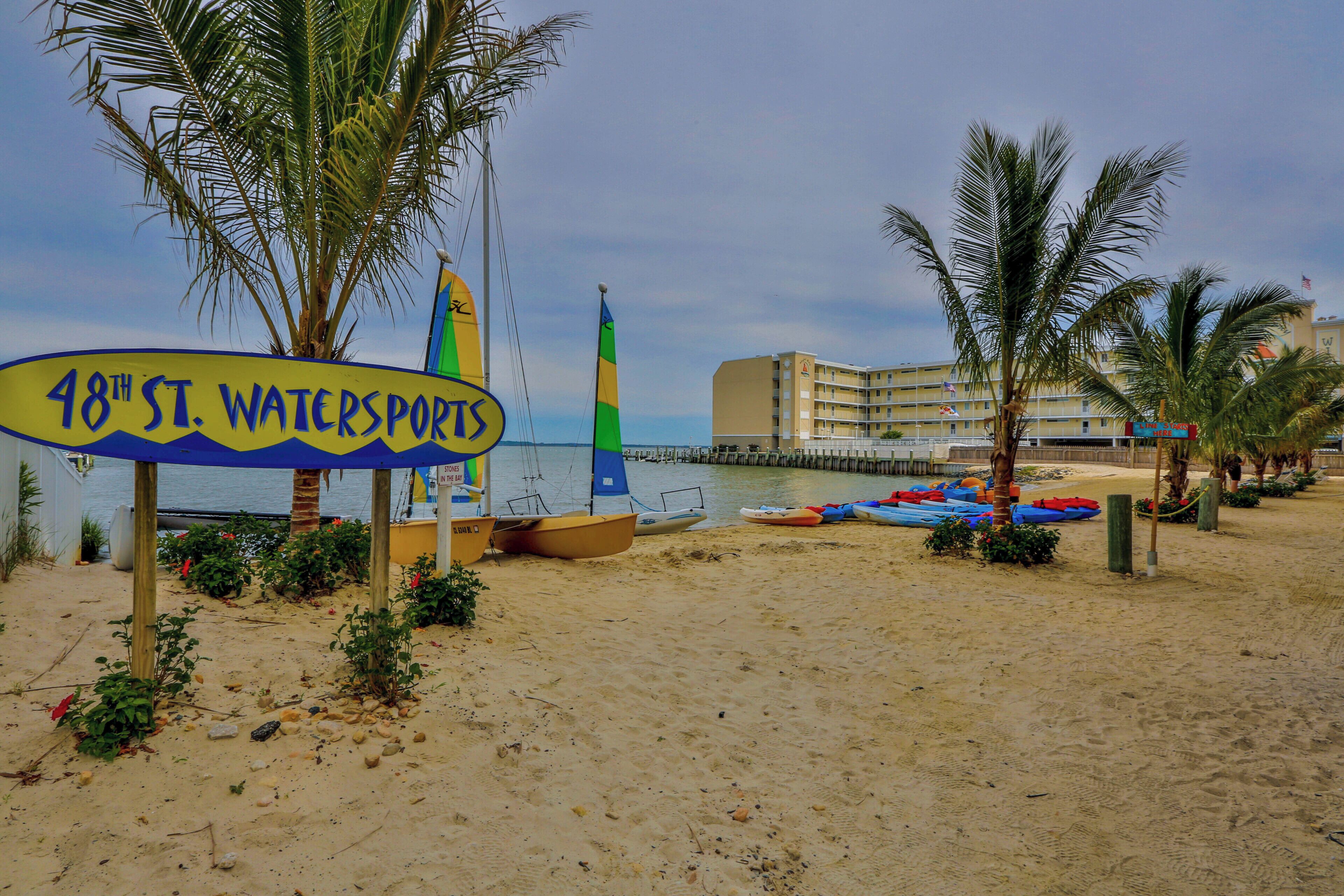 A beach with a sign for '48th St. Watersports,' boats, and a building in the background.