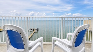 Two white chairs with blue cushions on a balcony with a white railing, overlooking calm water and a dock. - Princess Bayside Beach Hotel (Ocean City)